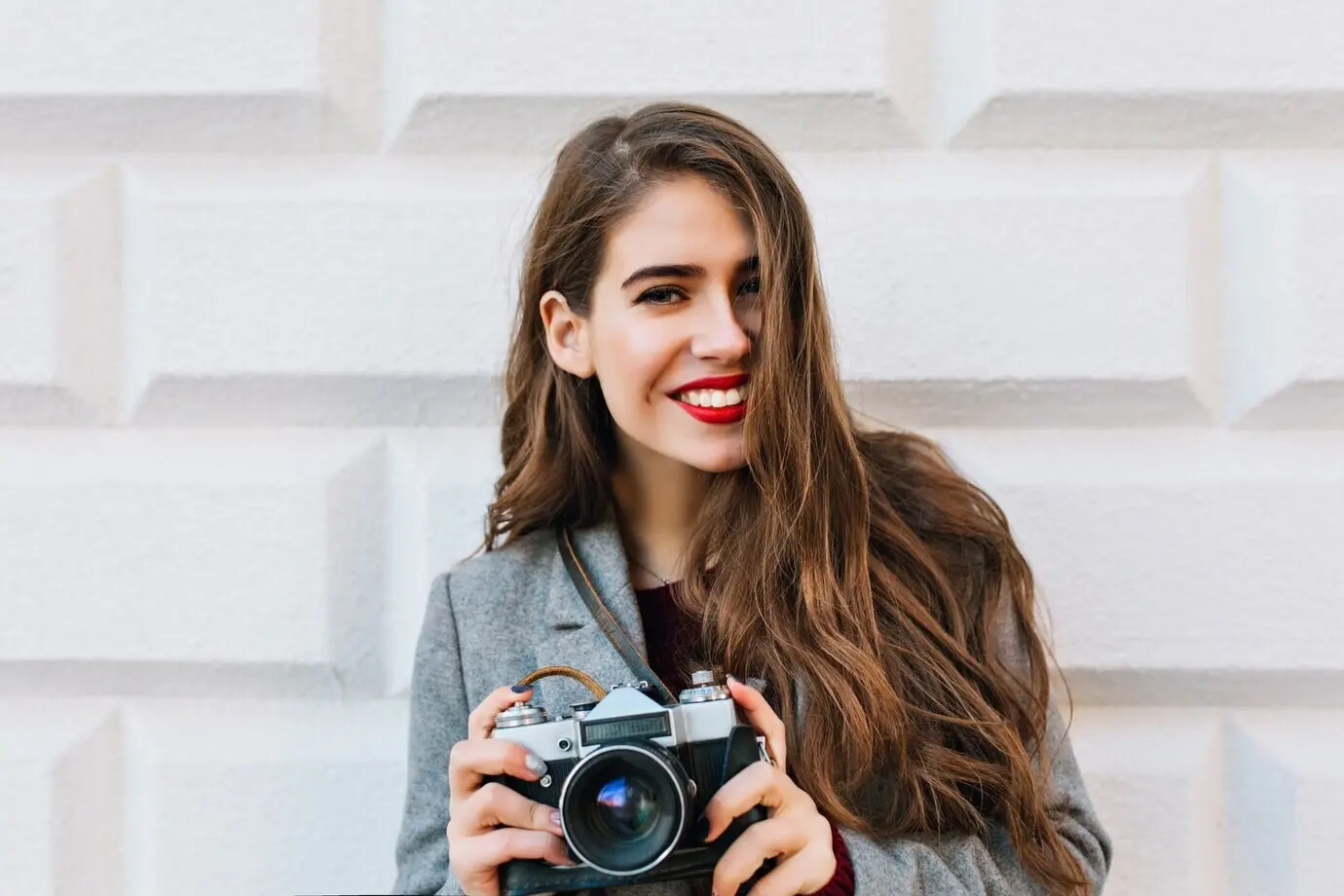 Porträt eines schönen Mädchens mit langen Haaren in grauem Mantel an einer Wand. Sie hält eine Kamera und sieht glücklich aus.